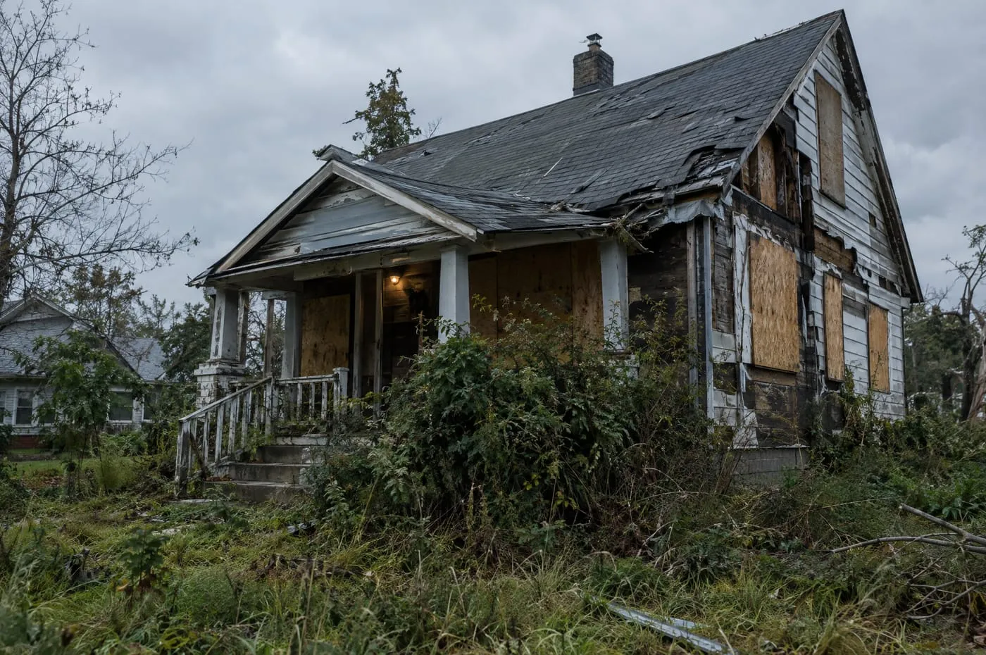 A boarded-up, storm-damaged house with a sagging roof and an overgrown yard.