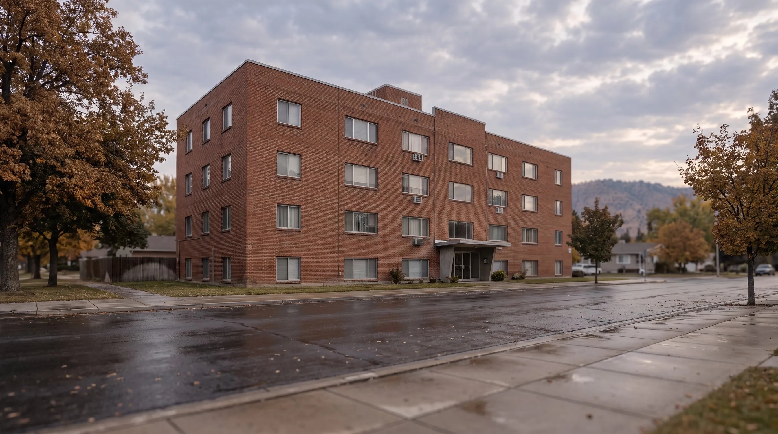 A four-story brick apartment building with a canopy entrance on a wet street.
