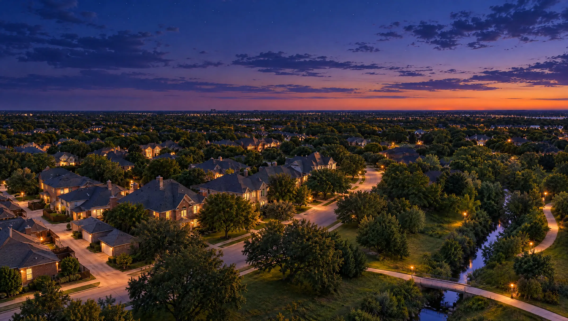 The same neighborhood from the air at dusk, with windows and streetlights lit and the creek reflecting the evening sky.