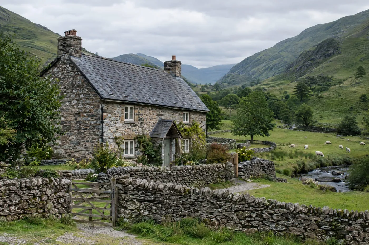 A stone holiday cottage with a slate roof in a green valley crossed by dry stone walls.