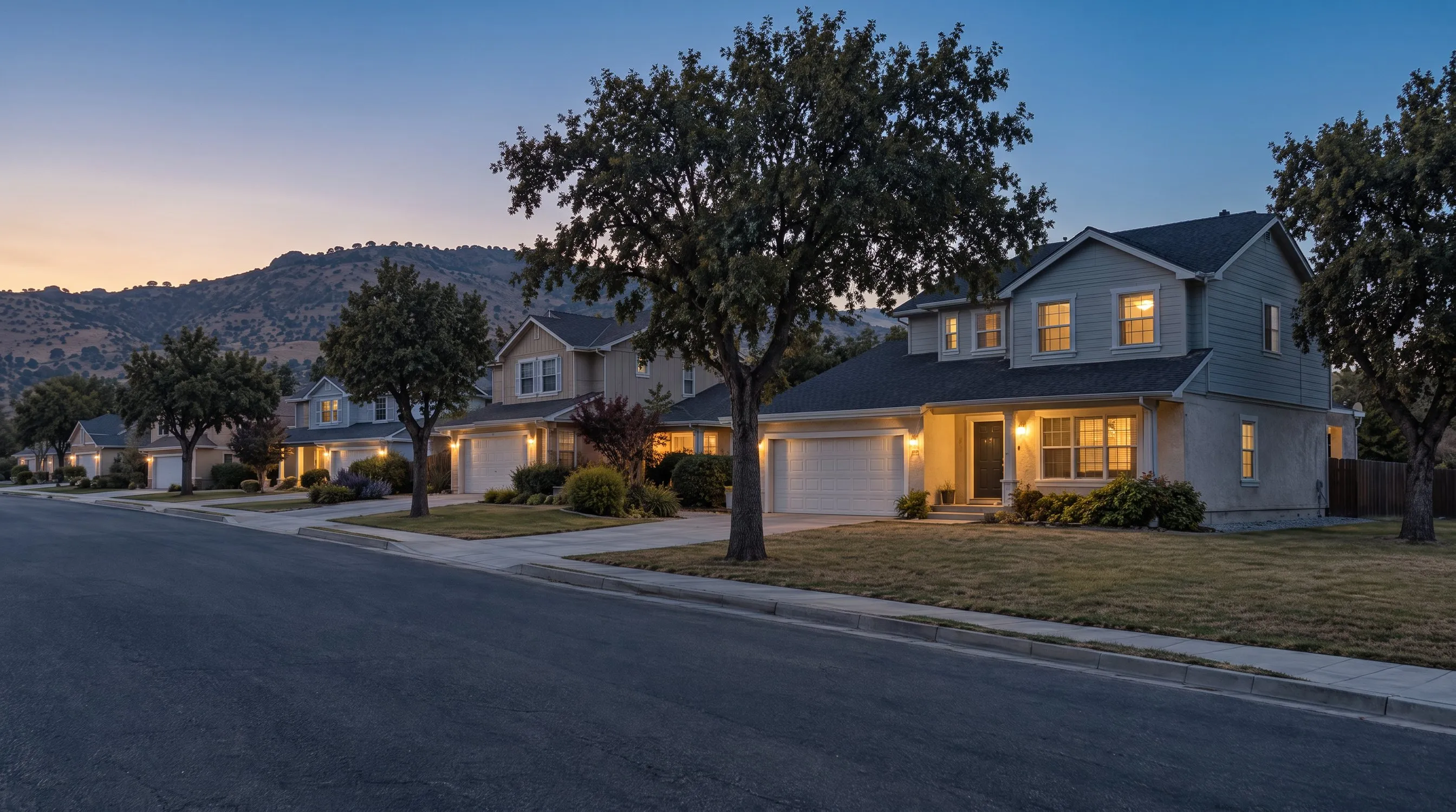 The same street at dusk, porch lights and windows lit, the ridge dark against a deep blue sky.