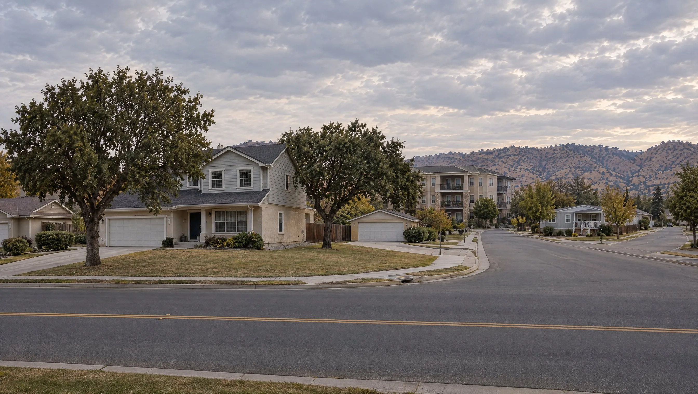 Wide view of the same street: the two-story house on its corner lot in front, a condominium building and a manufactured home farther down the road, a dry ridge behind.