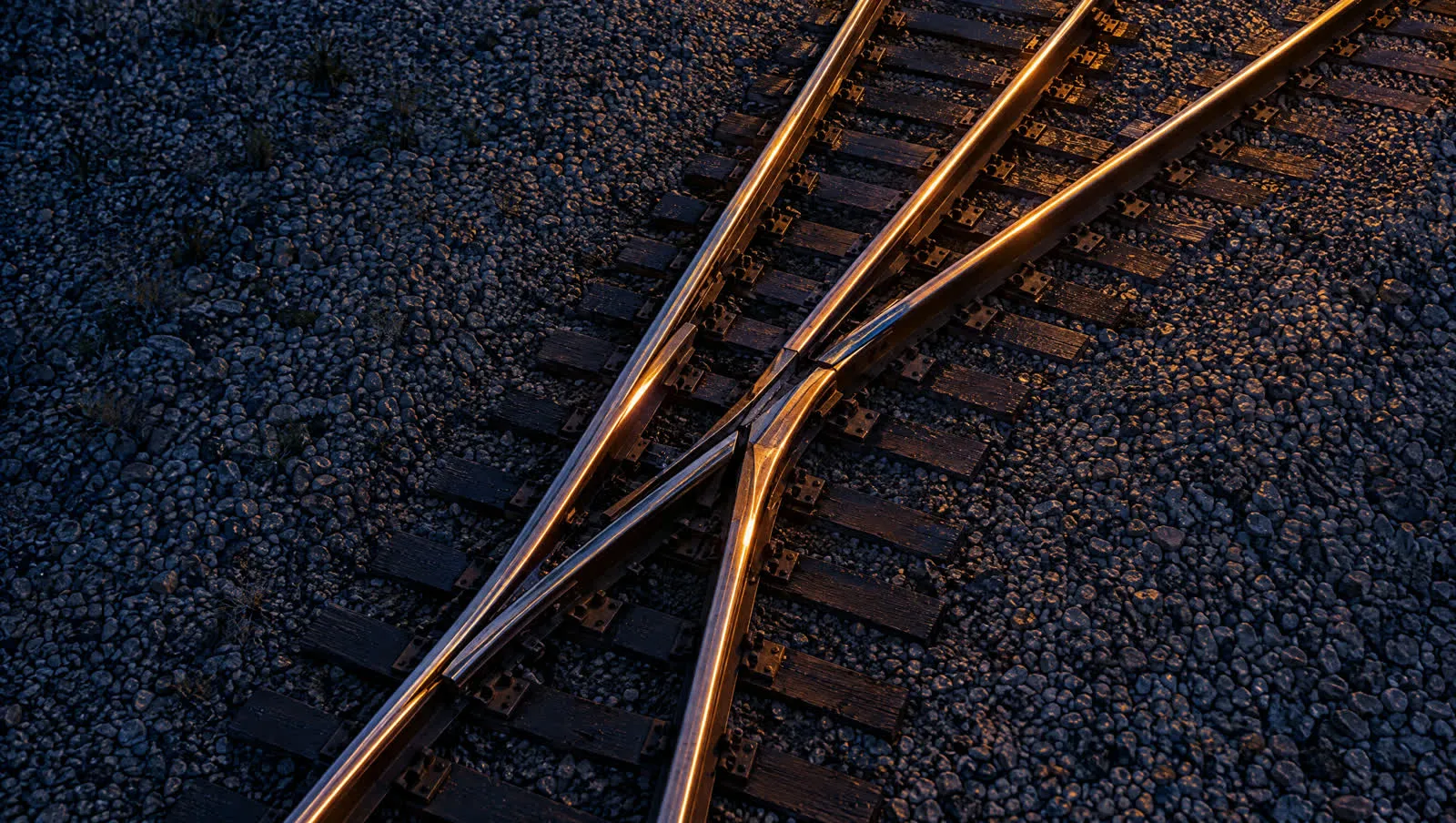 Overhead view of a railroad switch at night, where one track divides into two, with wet steel rails catching yard light.